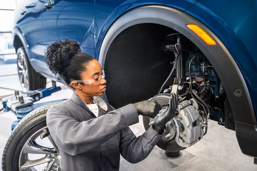 A mechanic with safety glasses and gloves works on a car's exposed front wheel hub in a garage.