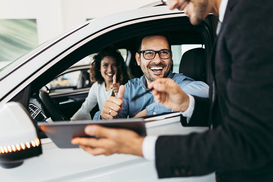 A couple giving thumbs up from inside a white car while a salesperson shows them documents on a tablet.