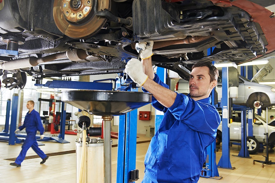 A mechanic in a blue uniform and white gloves works on the underside of a car hoisted on a lift. Another mechanic walks in the background.
